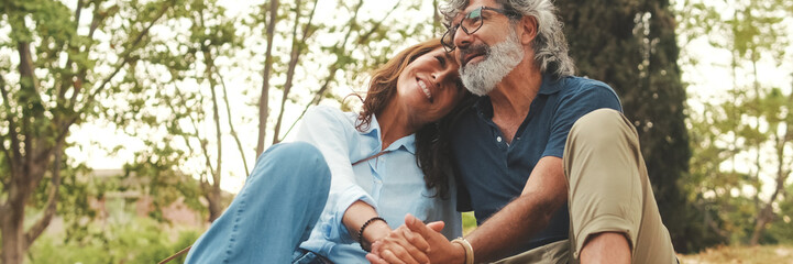 Fototapeta premium Happy retired couple sitting in the park holding hands and smiling resting on the grass in the park in autumn. Panoramic. Panoramic