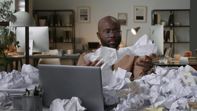 Zoom in shot of frustrated African American man covering his face with his hand amid chaos of crumpled papers on desk in office