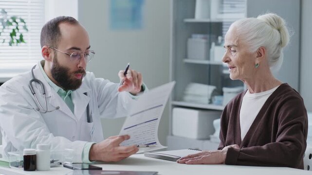 Professional male doctor reviewing medical records on clipboard and giving consultation to elderly female patient in clinic, discussing treatment plan and healthcare recommendations