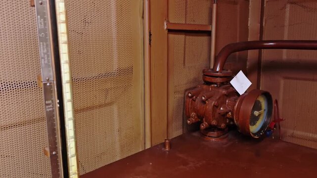 Rusted pump in metal compartment, brass gauge and vertical level stick visible inspection tag attached near flange, closeup of corroded surface in confined engine room, warm reddish industrial