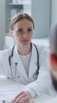 Female doctor sitting at desk with medical form on clipboard, discussing treatment with patient during consultation in clinic. Vertical format footage