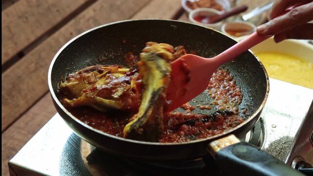 Close-up of pieces of fish being saut&eacute;ed in a rich, spicy red gravy in a non-stick pan. A person uses a pink silicone spatula to gently flip and coat the fish with the bubbling masala. The scene captu