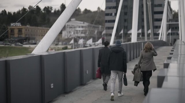 Three young business associates walk across a modern pedestrian bridge. The group moves together in an urban setting, reflecting teamwork and urban lifestyle.