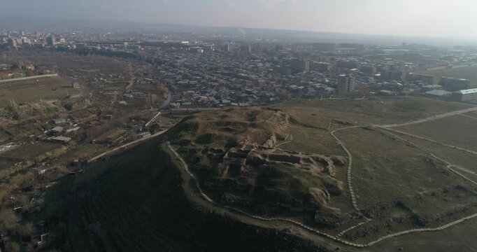 Breathtaking aerial shot of the ancient Erebuni Fortress ruins situated on a prominent hill. The drone flies forward revealing the archaeological site with the vast cityscape of Yerevan in the backgro