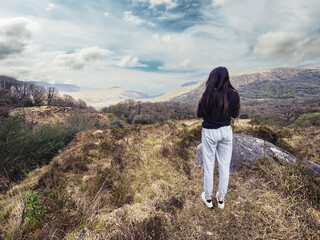 Woman stands alone on rugged hilltop overlooking vast, misty valley. Clouds drift above distant mountains, framing serene horizon. Her silhouette contrasts with earthy tones of grass and stone © mark_gusev