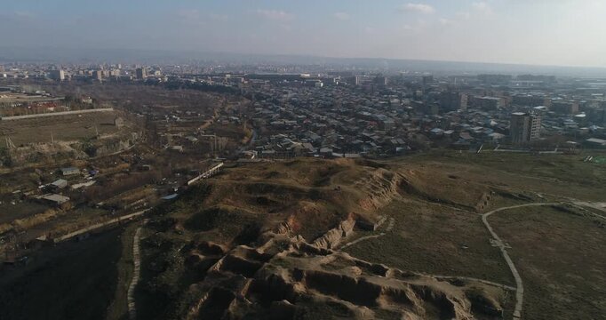 Breathtaking aerial shot of the ancient Erebuni Fortress ruins situated on a prominent hill. The drone flies forward revealing the archaeological site with the vast cityscape of Yerevan in the backgro