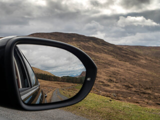 Reflection shows winding road through rugged hills. Cloudy sky looms over barren, rolling terrain. Car side mirror frames the journey ahead. Driver's presence implied, yet unseen. Wild Ireland nature