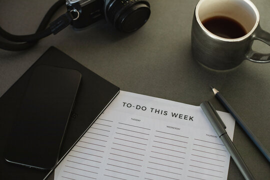 Close-up of a To do list, notebook, pens, mobile phone, camera and a cup of black coffee on a desk