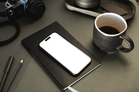 Close-up of a mobile phone with a blank screen on a notebook with a cup of black coffee and camera on a desk