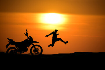 Silhouette of a man leaping near a motorcycle silhouetted against an orange sunset