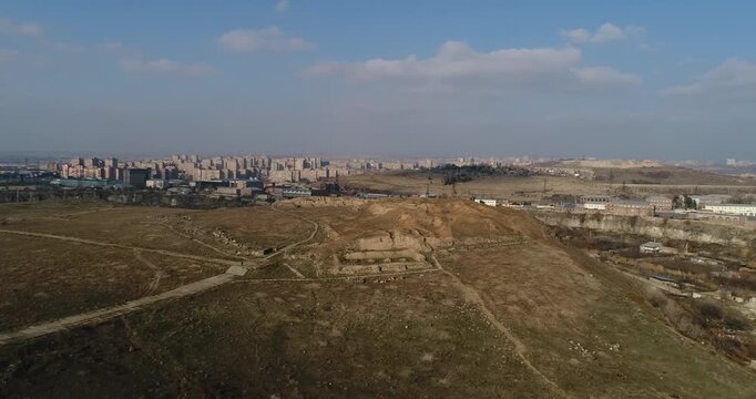 Breathtaking aerial shot of the ancient Erebuni Fortress ruins situated on a prominent hill. The drone flies forward revealing the archaeological site with the vast cityscape of Yerevan in the backgro