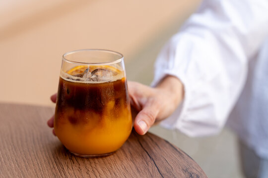 Close-up of a woman's hand reaching for a glass of orange Espresso Tonic with Ice on a wooden table