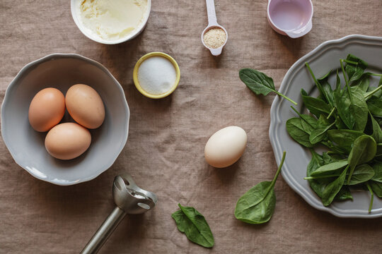 Overhead view of healthy gluten free pancake ingredients and kitchen utensils including eggs, fresh spinach, mascarpone cheese, salt, and psyllium on a linen tablecloth