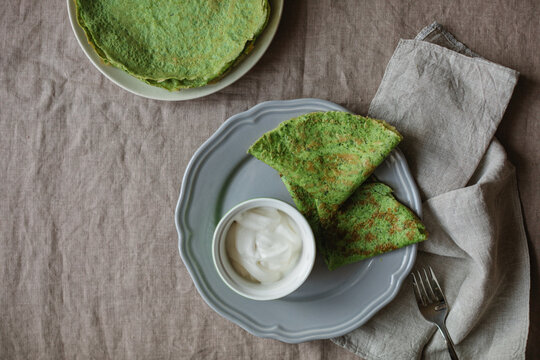 Overhead view of a stack of homemade gluten free spinach pancakes, two pancakes on a plate with a bowl of greek yogurt on a linen tablecloth with a napkin