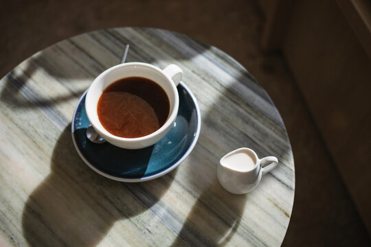 Overhead view of a cup of black coffee and a jug of milk on a marble table in sunlight