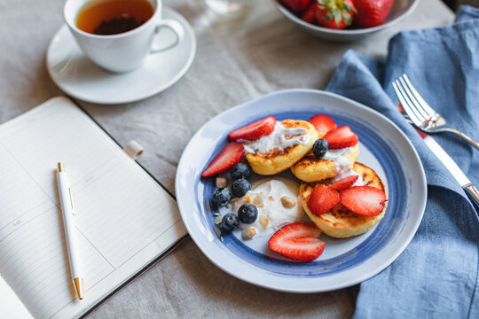 Overhead view of homemade keto cottage cheese pancakes with fresh strawberries and blueberries, a bowl of strawberries and a cup of tea on a table with an open notebook and pen
