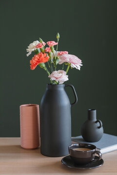 Cup of coffee, a bunch of pink and orange carnation and ranunculus flowers, assorted vases and a notebook on a table