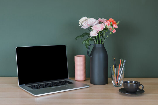 Laptop computer with a blank screen, a cup of coffee, a bunch of pink and orange carnation and ranunculus flowers in a ceramic vase and pens in a glass on a desk