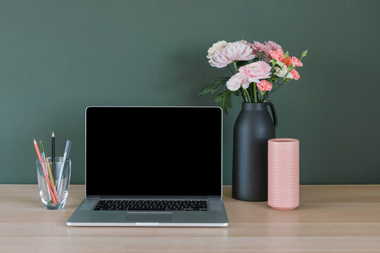 Laptop computer with a blank screen, a bunch of pink and orange carnation and ranunculus flowers in a ceramic vase and pens in a glass on a desk