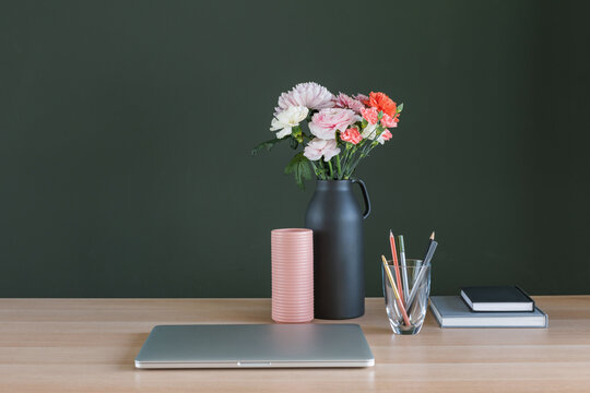 Laptop computer, a bunch of pink and orange carnation and ranunculus flowers in a ceramic vase, pens and notepads on a desk