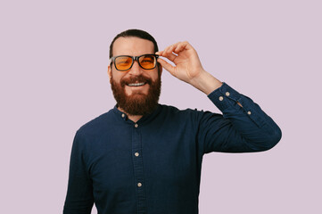 Obraz premium Bearded Man Adjusting Glasses Playful Smile, Studio Headshot On Lavender Background, Amber Tinted Frames,