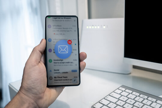 Closeup of male hand holding smartphone displaying email inbox interface with red notification bubble icon of unread messages in white home office with computer keyboard and wifi internet router