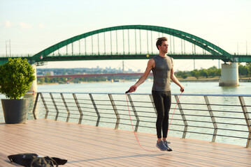 Naklejka premium Male athlete skipping rope for fitness and cardio workout on a riverside pier with a green arch bridge spanning the river in a city during daylight