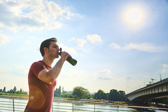 Young athletic man drinking water outdoors, staying hydrated and refreshed during a hot summer day, with a river and bridge visible in the urban landscape
