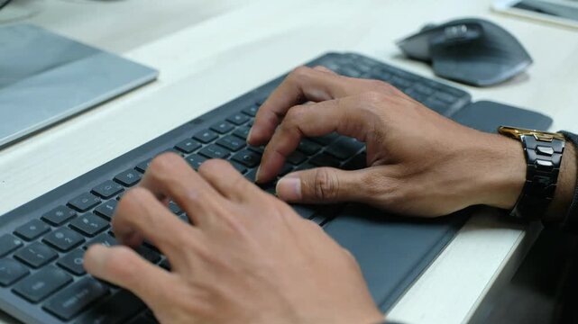 High-quality close-up footage of a man's hands typing rapidly on a modern black wireless keyboard at a wooden desk.