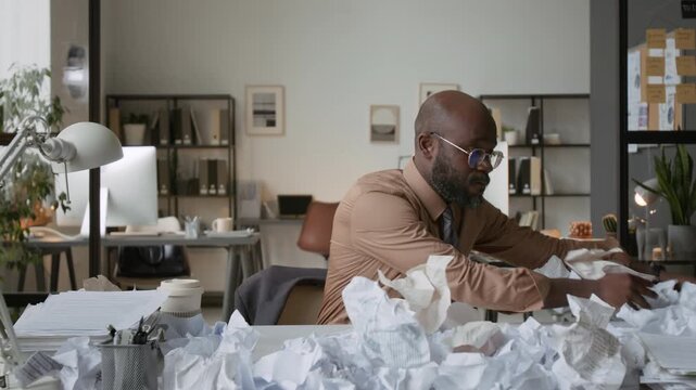 Zoom in shot of African American man sweeping huge pile of crumpled sheets of paper off his office desk and putting his head on it in despair