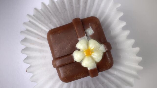 Top view of square milk chocolate praline with white sugar flower in paper cup. Artisan confectionery on white background. Gourmet sweet dessert, luxury handmade candy concept.
