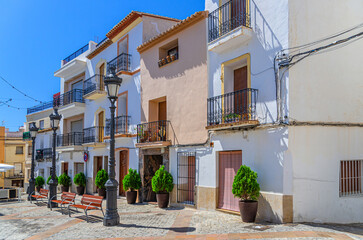 Obraz premium Old town Calpe city historic centre with white houses traditional buildings, benches and street lights on small square Villa Plaza in sunny summer day, Calp Casco Antiguo, Valencian Community, Spain