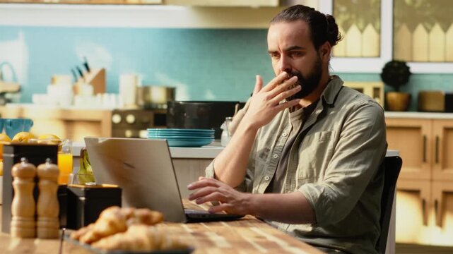Uncertain person dealing with ambiguous remote job tasks at home, preoccupied with misunderstanding and disorganized data. Confused unsure man, frowning and struggling at kitchen table.