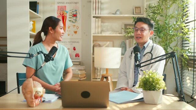 Asian medical doctor hosting a healthcare podcast interview with a nurse inside a bright clinic office. Professional consultation recording with microphones and laptop discussing treatment, patient	
