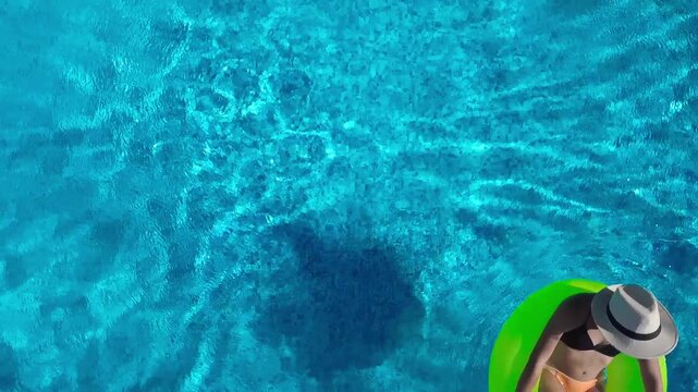 Aerial drone view of young woman in hat relaxing in swimming pool, top view of beautiful girl in bikini floats on inflatable ring donut and has fun in water, tropical vacation on holiday resort