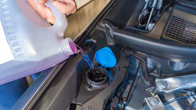 Person pouring windshield washer fluid from a container into a car's reservoir under the hood, showcasing automotive maintenance in a garage setting