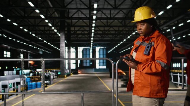 Black female worker evaluates heavy equipment activity on tablet, in a busy engineering plant. Machinery hall filled with steel work, welding activity and mechanical systems for heavy industry.