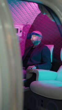 Relaxed man wearing a virtual reality headset while sitting in a futuristic cabin during a chromotherapy and hyperbaric oxygen therapy session in a modern wellness spa for mental health