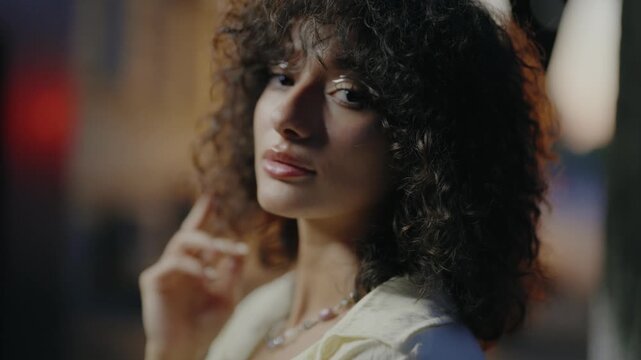 Stunning young woman with voluminous curly hair and glamorous makeup posing sensually and looking at the camera in a dimly lit, atmospheric setting with soft, blurred background lights