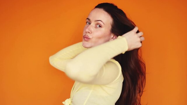 Happy young woman touching long brunette hair, turning in profile and pouting lips on orange background. Beautiful girl posing, looking up and smiling at camera. Haircare and beauty concept.
