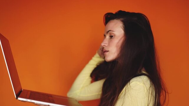 Side view of serious brunette woman working on laptop and typing on orange background. Concentrated girl using silver computer, scrolling and checking information. Freelance and study concept