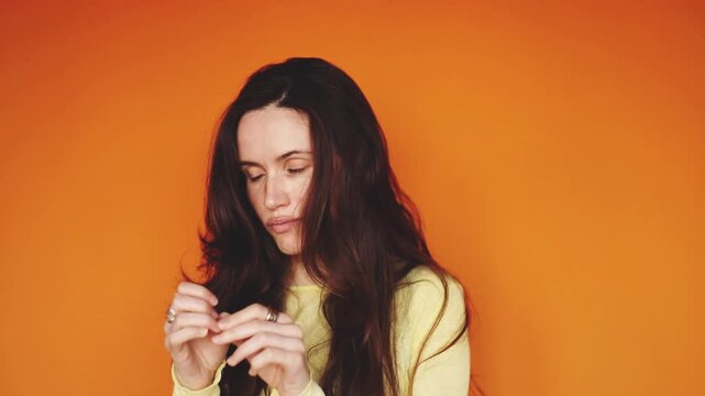 Horizontal video of arrogant brunette woman looking at her nails with boredom on orange background. Haughty girl feeling annoyed and looking away with disdain. Superiority and negative emotions
