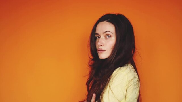 Horizontal video of arrogant brunette woman standing in profile and turning to camera with disdain. Haughty girl with crossed arms showing boredom and indifference on orange background. Ego concept