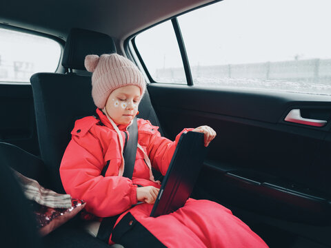 A girl child in a pink jumpsuit is fastened with a seat belt in the car with a tablet in her hands.