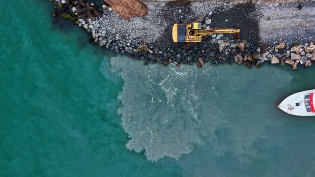 Heavy machinery operates at the edge of the water, mixing soil and sediments on the shore under clear skies. Waves lap gently on the rocks and sand.