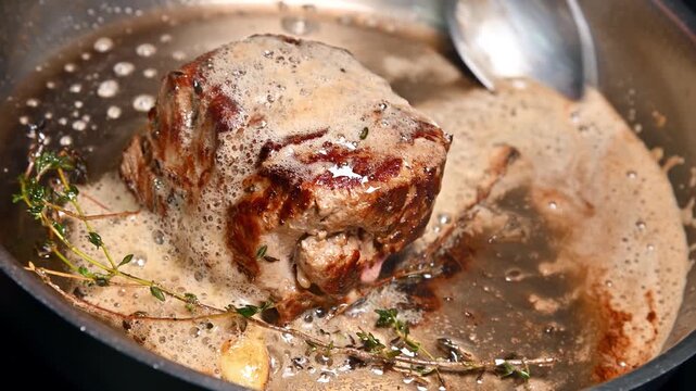 Chef seasons and bastes meat in pan with herbs and bubbling sauce