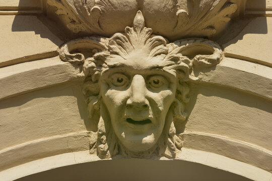 A close up of a decorative stone mascaron depicting a human face with expressive features and acanthus leaf ornaments on a classic building facade.