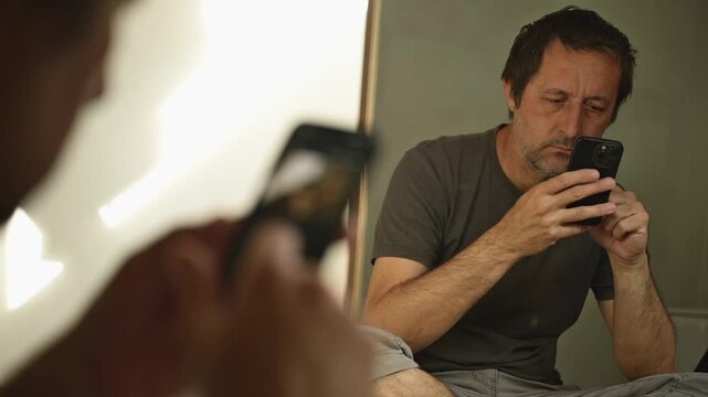 Serious mature man using smartphone while sitting in front of a large mirror in his living room. 4K