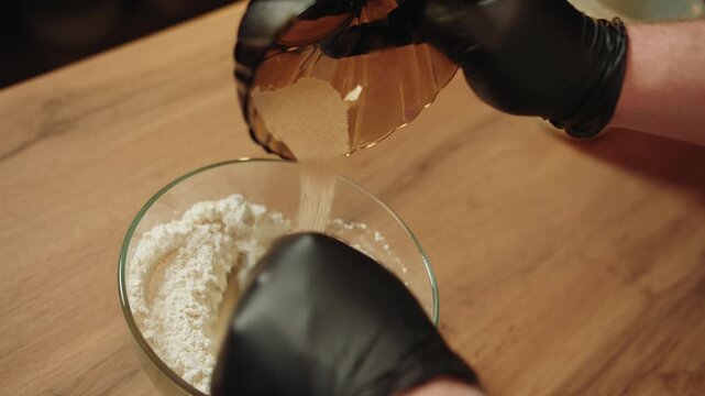 Table top view of male hands in black gloves that pour from small bowl the dry yeast into larger glass bowl with wheat flour. Closeup of mixing ingredients to make dough on wooden table. No face