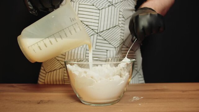 Man in apron pours measured milk from plastic jug into flour in glass bowl. Hands in black latex gloves knead batter on wooden table. Stirring dough in kitchen and cooking at home